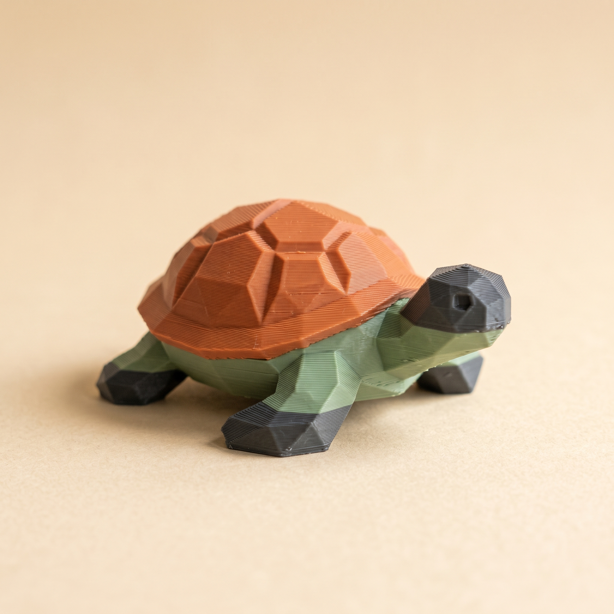A small multi-color FDM 3D-printed low-poly turtle figurine with a sage green body, terracotta orange shell sections, and charcoal grey feet on cream paper