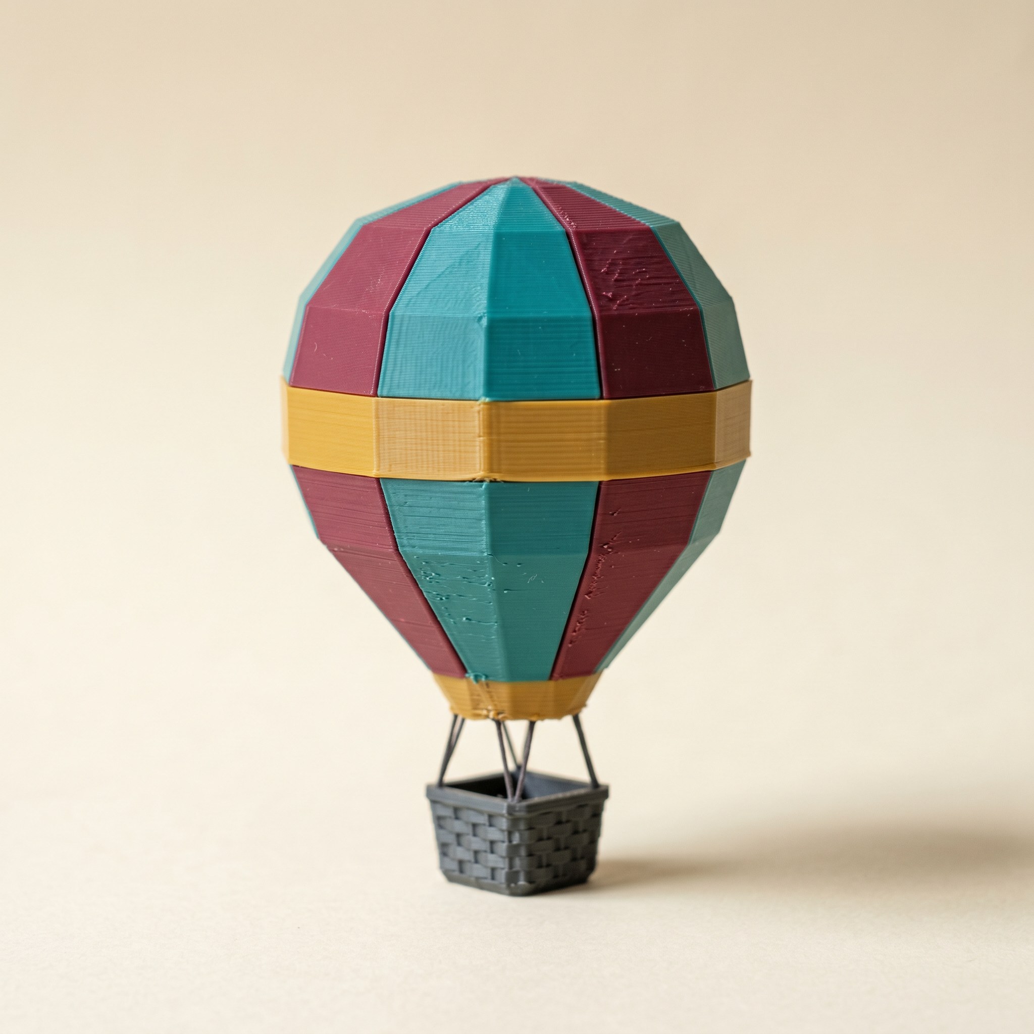 A low-poly multi-color FDM 3D-printed hot-air balloon in matte teal, burgundy, and mustard with a charcoal basket, on cream paper