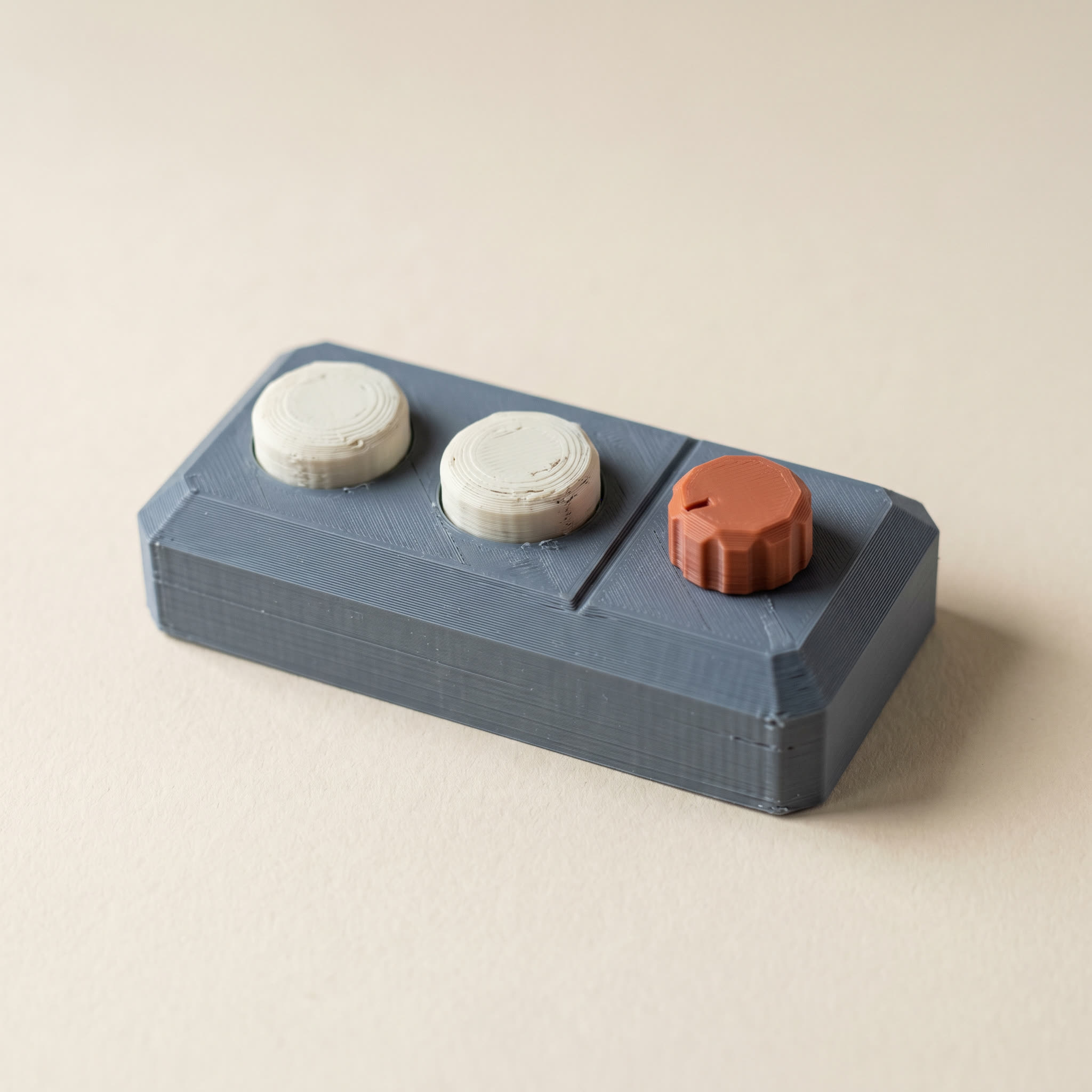 A small multi-color FDM 3D-printed low-poly sci-fi control panel block with matte slate-gray body, two raised cream button caps, and a small terracotta-orange dial on cream paper