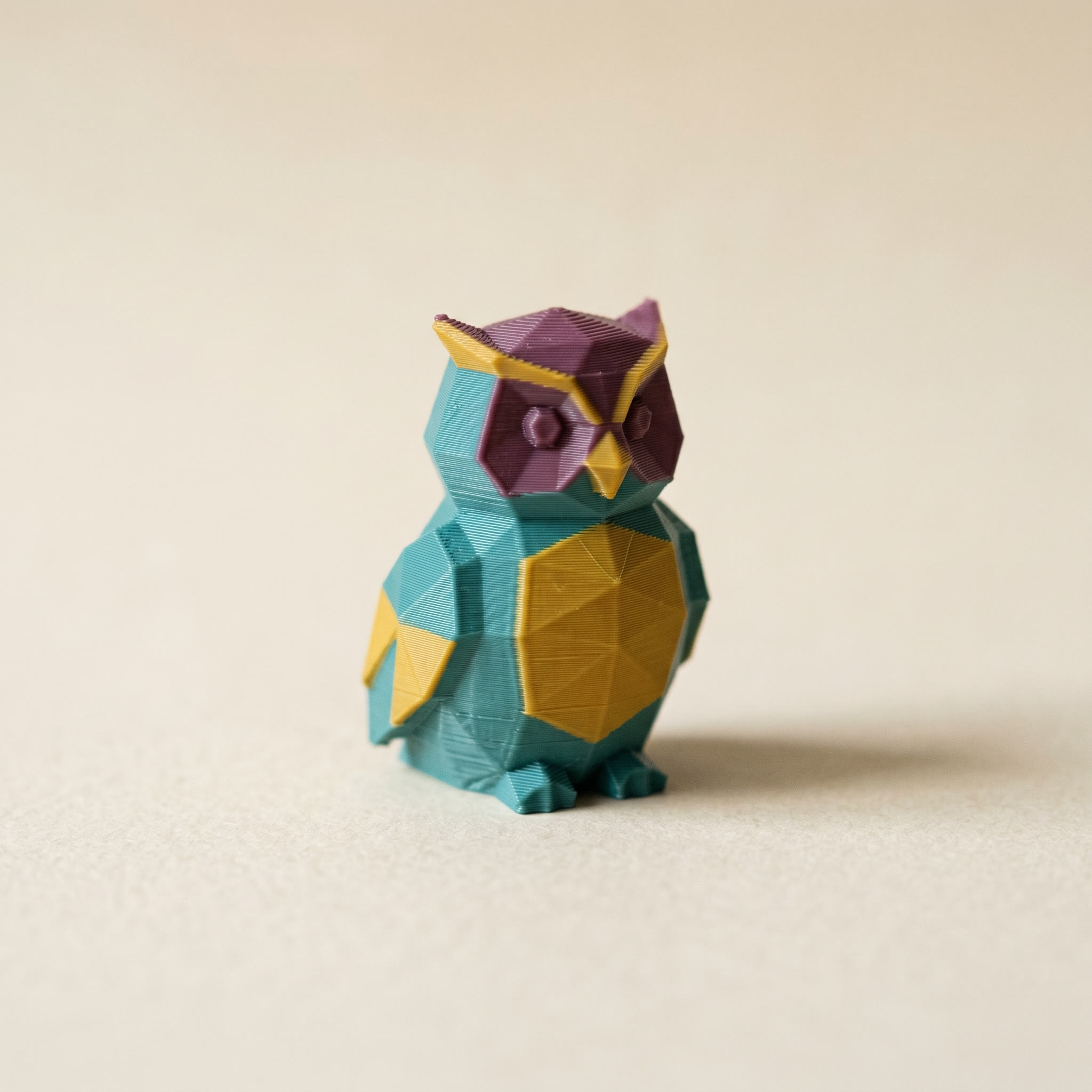 A small multi-color FDM 3D-printed low-poly owl figurine in matte teal, mustard yellow, and plum filament on cream paper