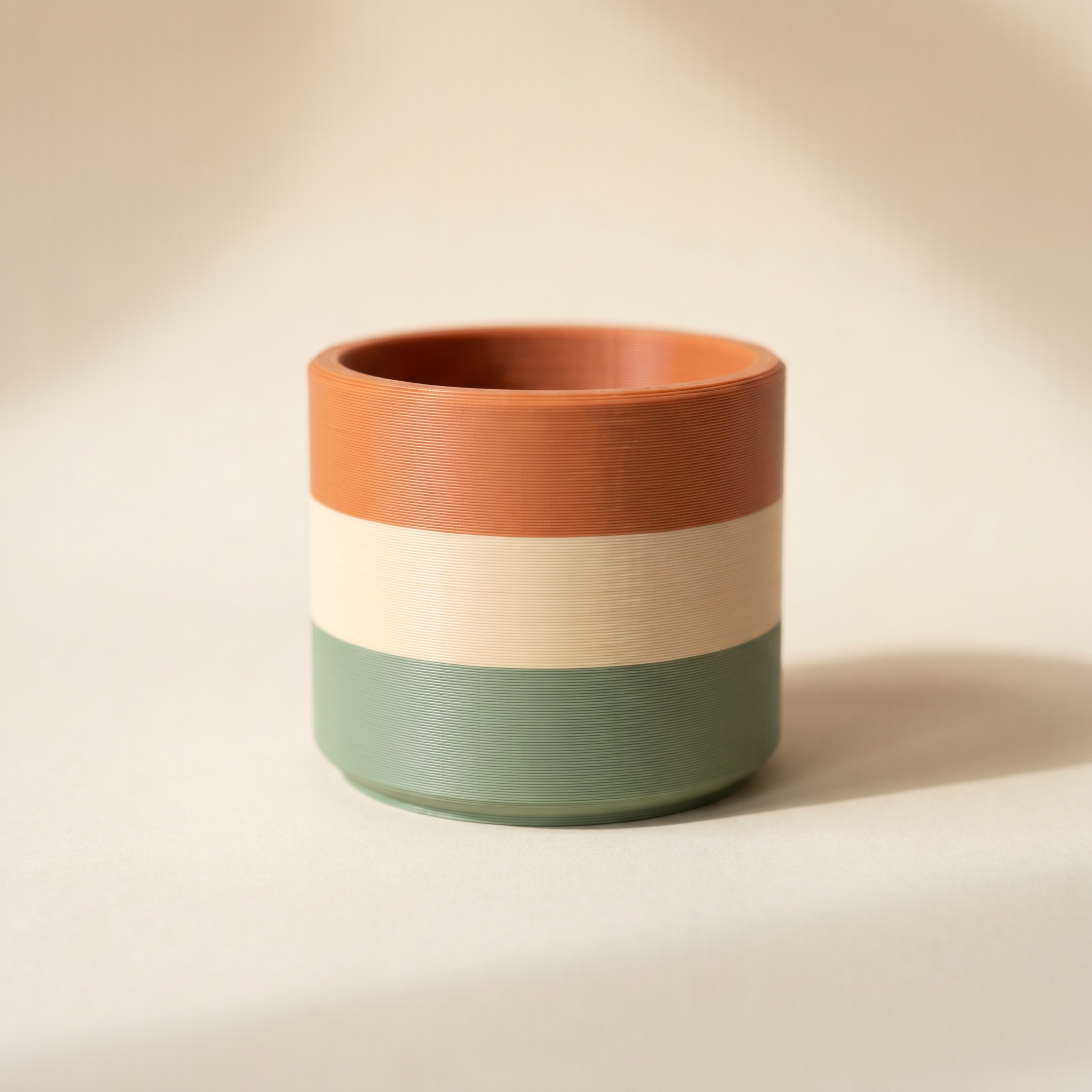 A multi-color FDM 3D-printed planter pot with horizontal sage green, cream, and terracotta filament bands on cream paper