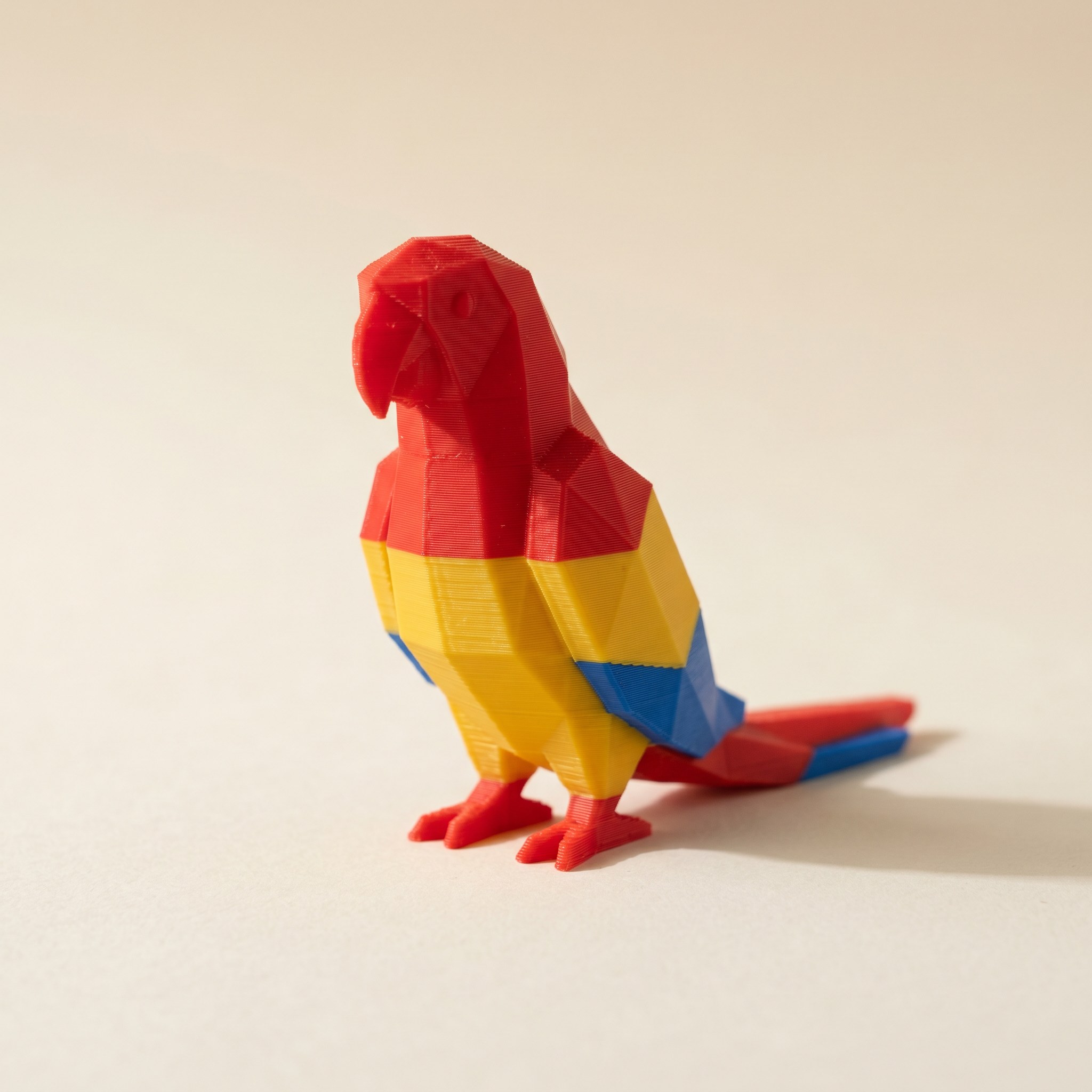 A small multi-color FDM 3D-printed low-poly macaw figurine in matte scarlet, golden yellow, and cobalt blue filament on cream paper
