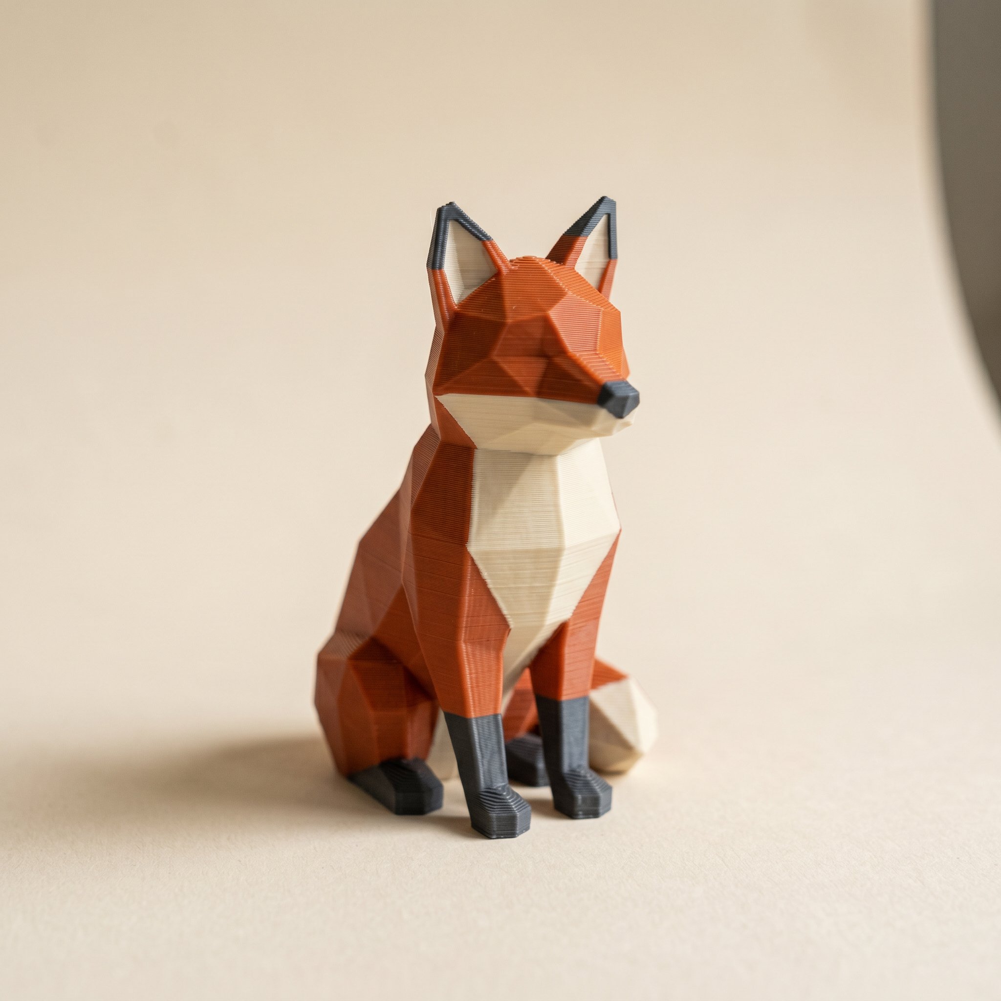 A low-poly fox 3D printed in matte burnt-orange, cream, and charcoal filament, showing clean color boundaries printed without paint