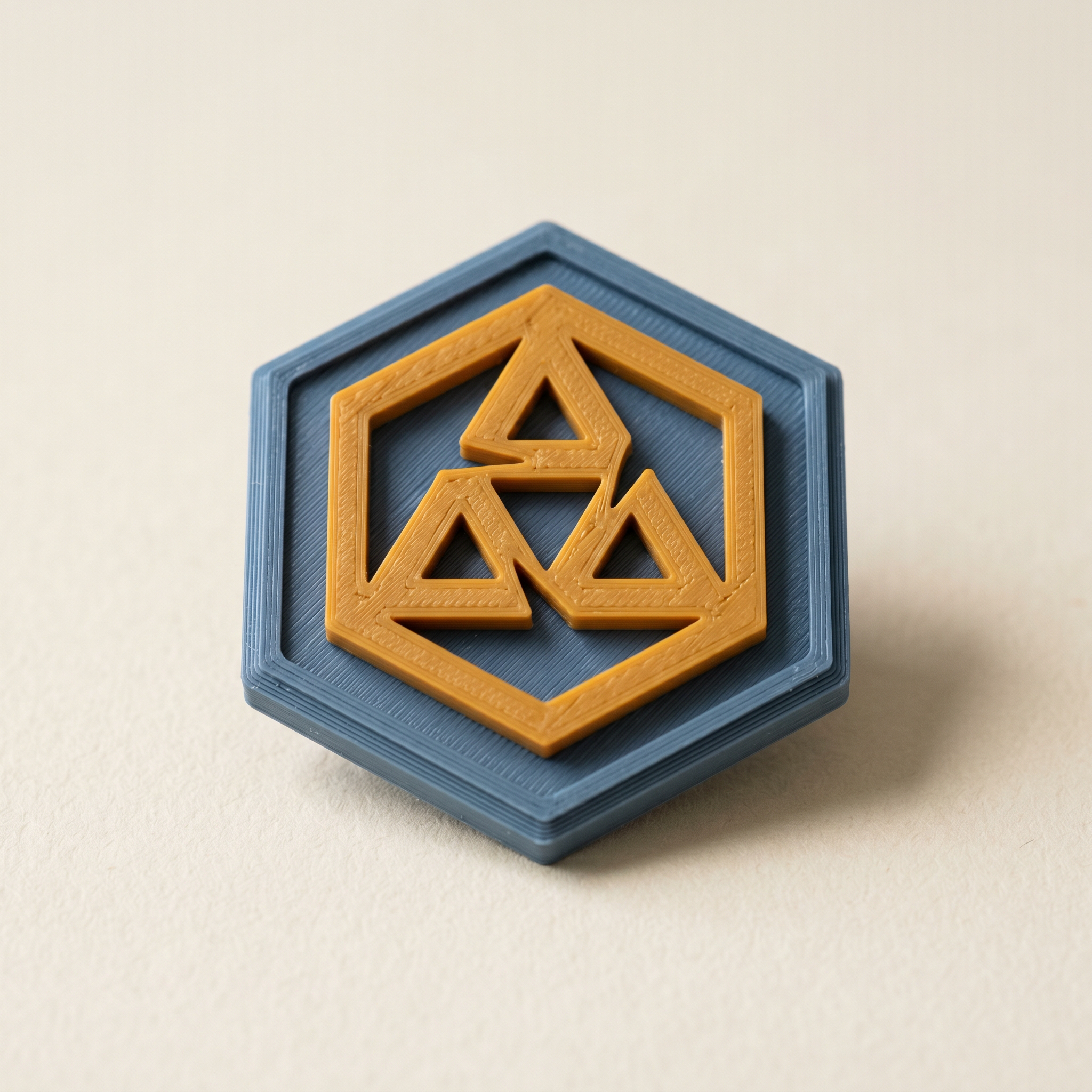 A multi-color FDM 3D-printed hexagonal badge with a raised mustard-orange emblem standing proud of a matte slate-blue backing plate, on cream paper