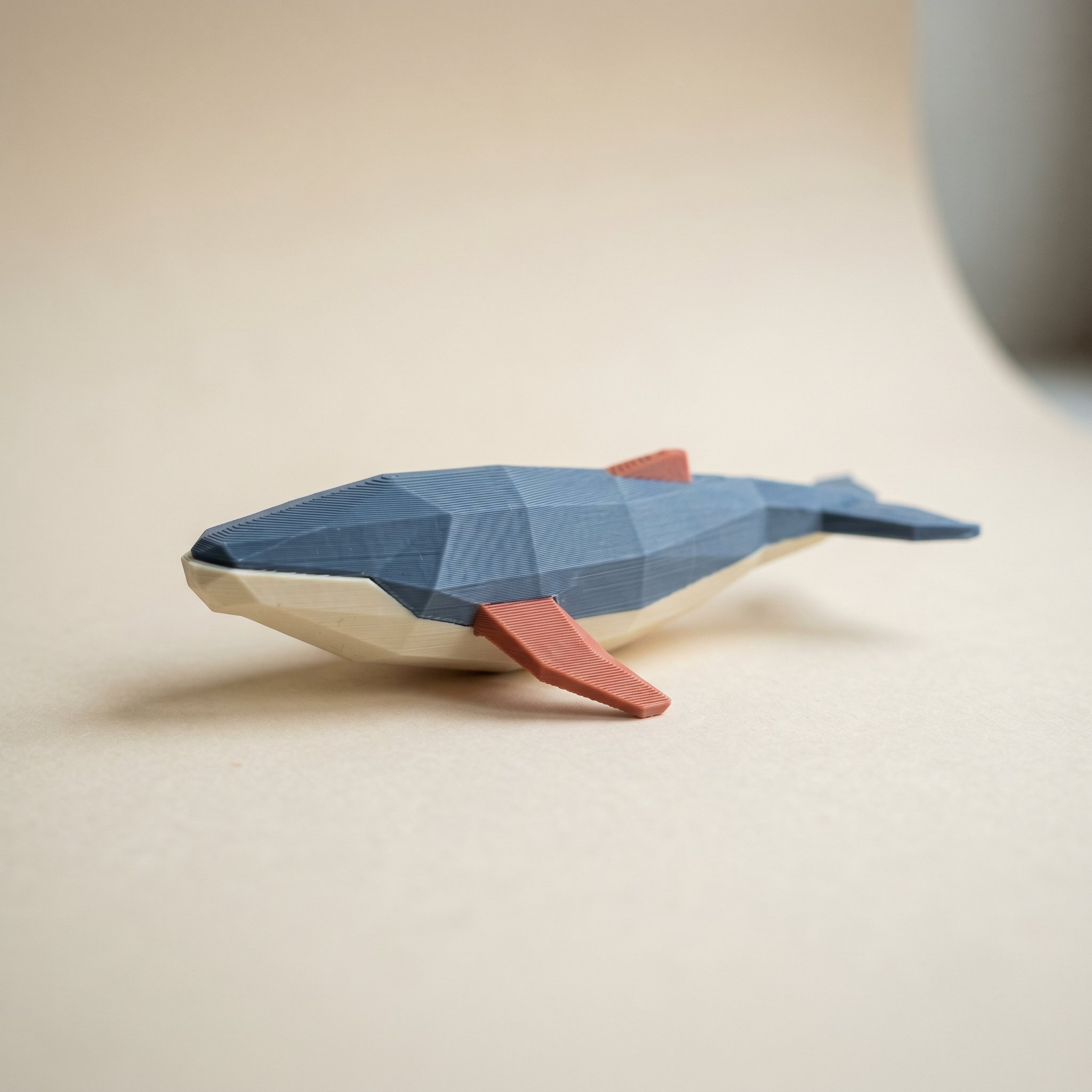 A multi-color 3D printed low-poly whale figurine in slate blue, coral, and cream filament, the kind of painted model shared as a MakerWorld print profile