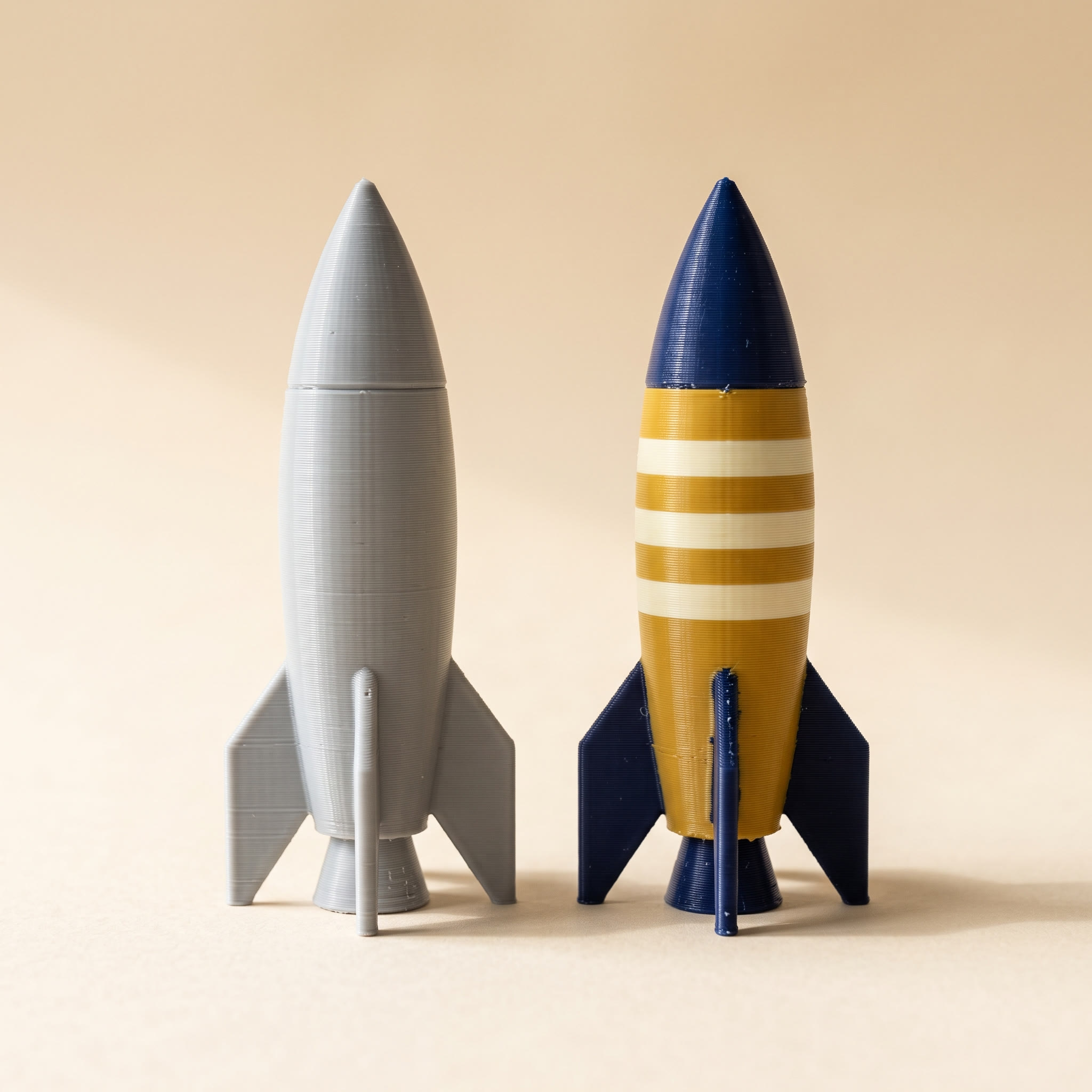 Two 3D-printed model rockets side by side, one in plain grey filament like an STL to OBJ conversion produces and one printed in navy, mustard and cream filament