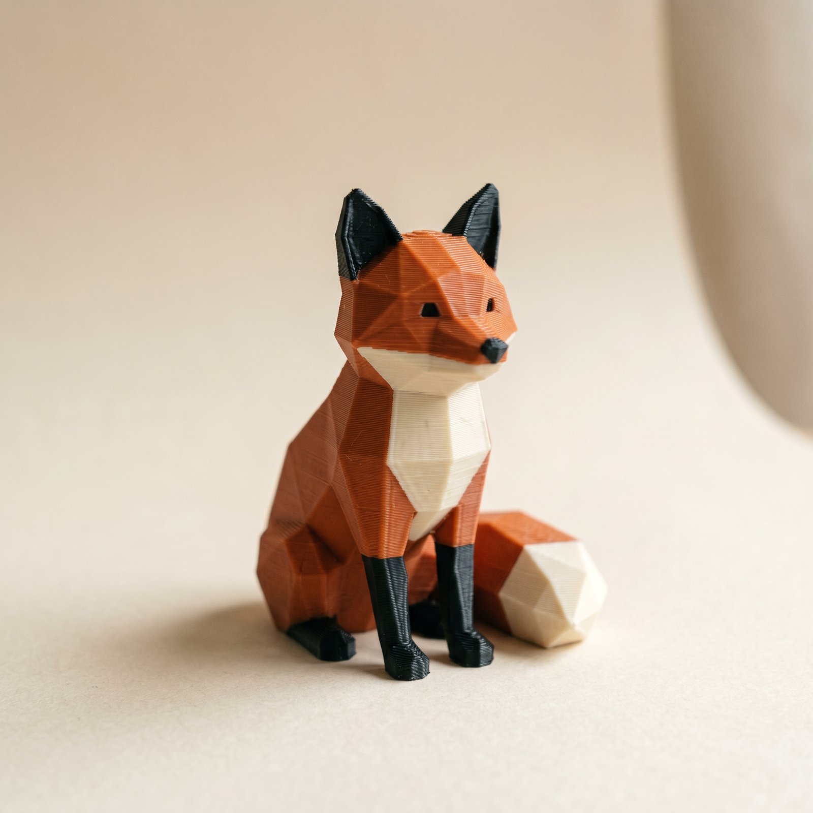 A small multi-color FDM 3D-printed low-poly fox figurine in matte burnt-orange, charcoal, and ivory filament on cream paper